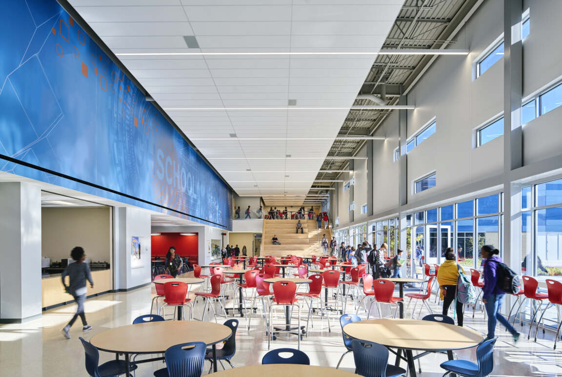Modern school cafeteria with red chairs, open seating, high ceilings, natural light, and students socializing.