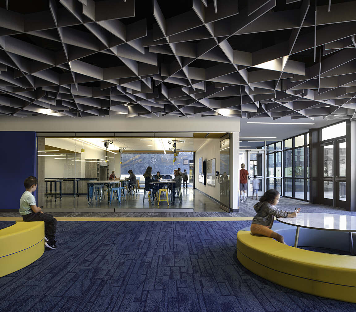 Modern school commons with geometric ceiling panels, blue carpeting, and curved seating areas, leading into a collaborative STEM classroom.