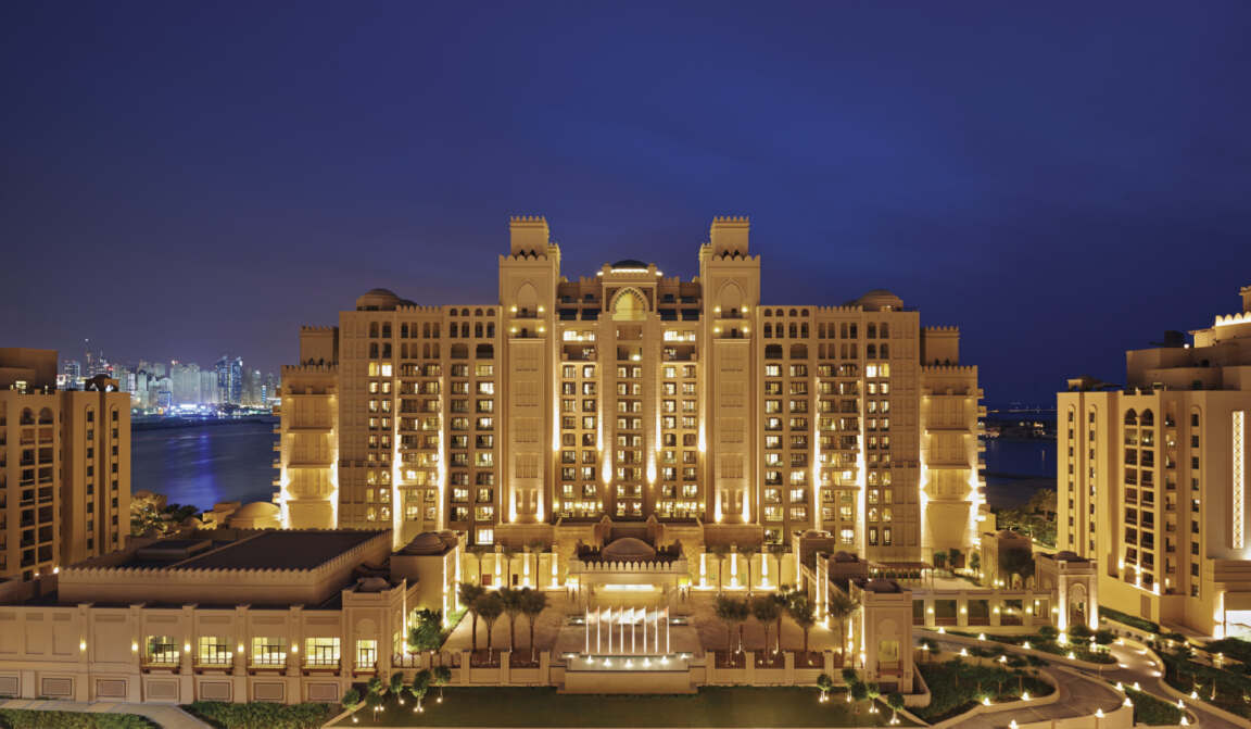 Luxury waterfront resort illuminated at night, showcasing grand architecture with glowing lights and a serene view of the water.