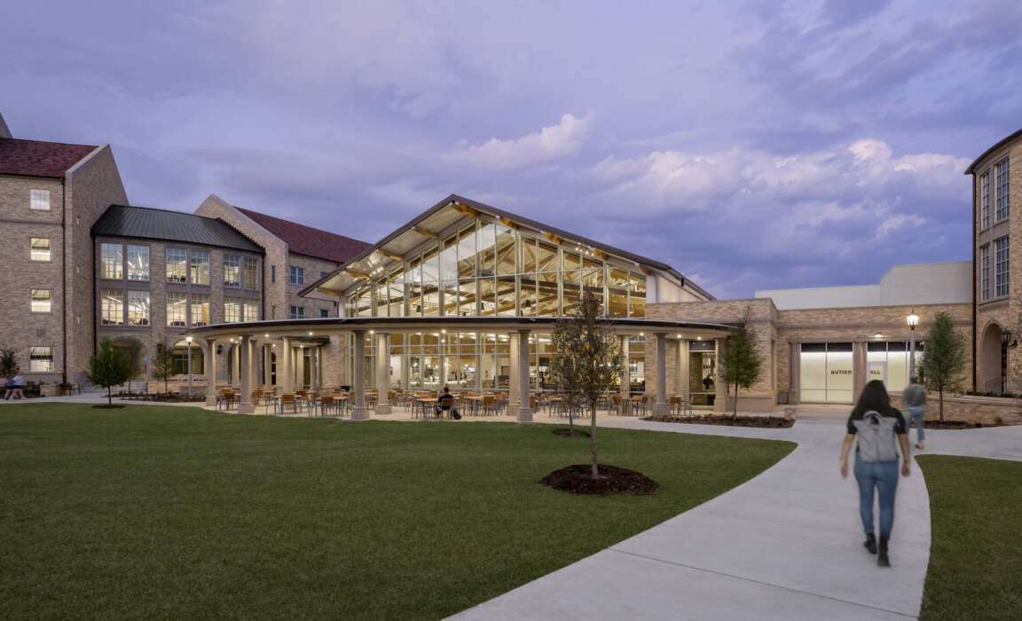 A modern glass dining hall with a high roof and outdoor patio is nestled between traditional brick buildings under a dramatic twilight sky.