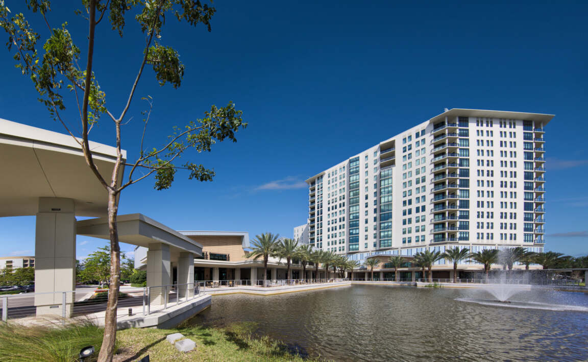 A towering residential building along a large pond with a fountain and neat landscaping.