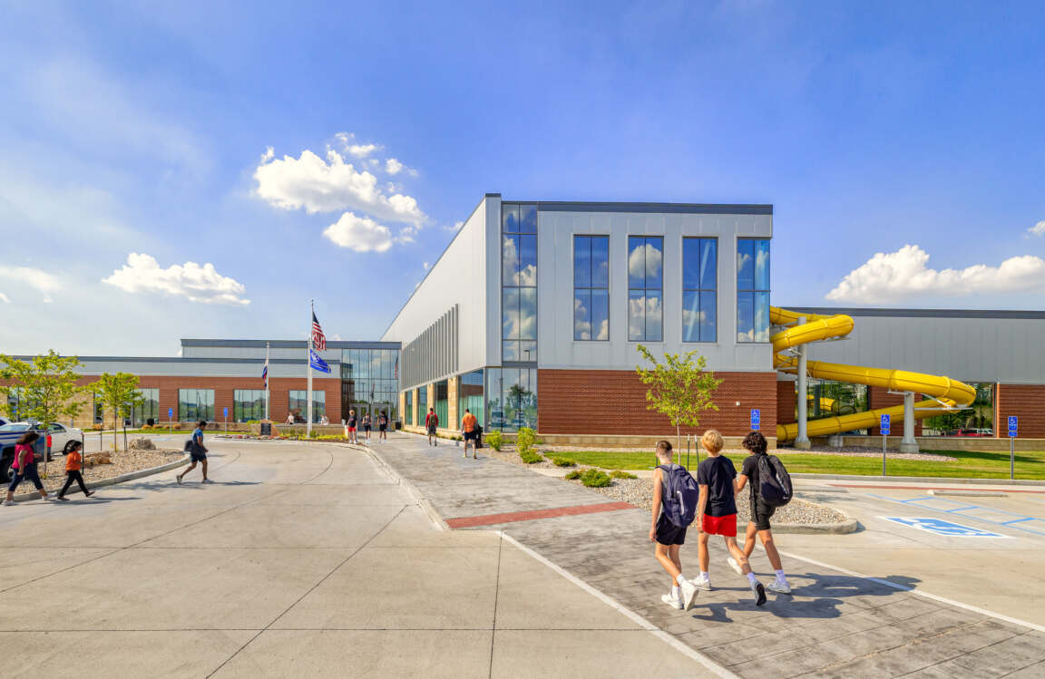 A bright, sunny shot of a modern building with red brick and a large windowed section. People walk by, and a yellow water slide is visible on the right.