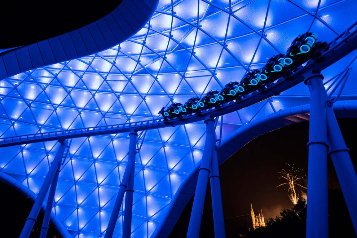 Futuristic roller coaster glides under a glowing blue canopy at night, blending dynamic architecture with thrilling motion and ambient lighting.