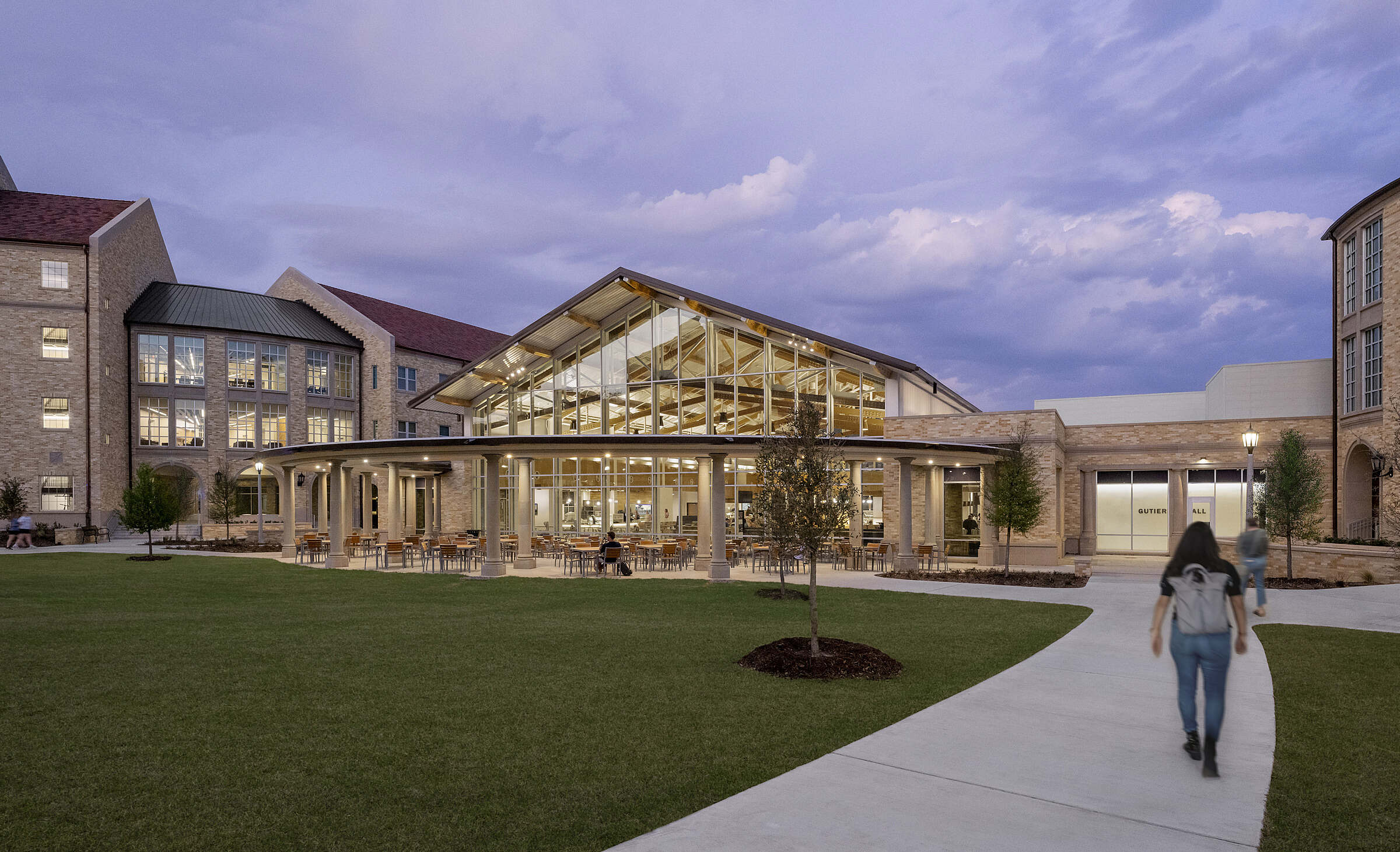 A modern glass dining hall with a high roof and outdoor patio is nestled between traditional brick buildings under a dramatic twilight sky.